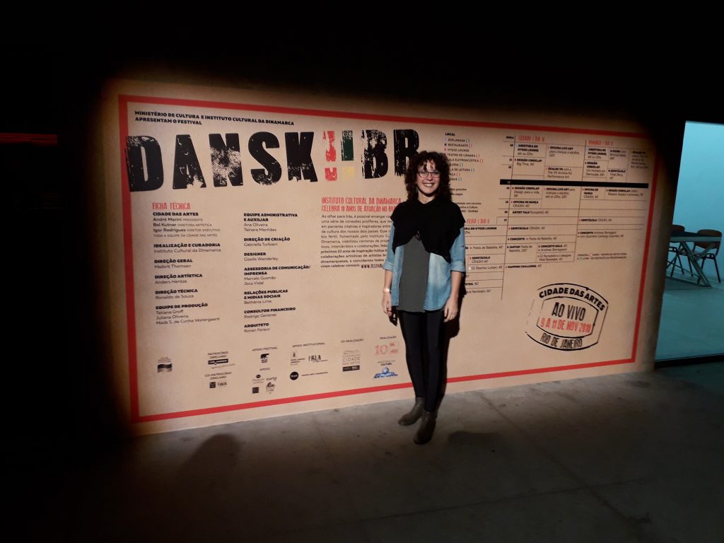 Gabriella Turbiani na frente do Banner Ficha técnica e programação do Festival Dansk!!BR